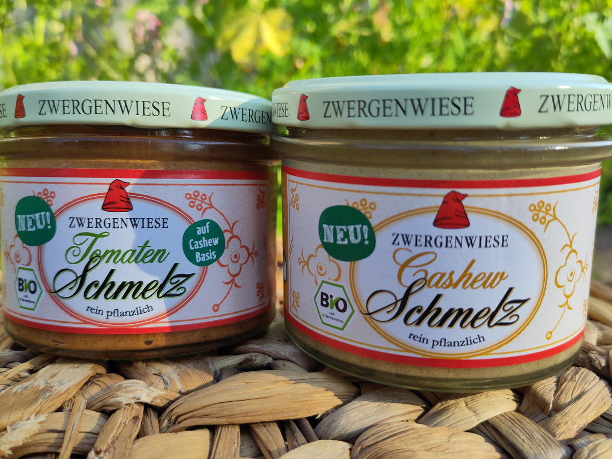 KI generiert: Zwei Zwergenwiese Gläser mit Tomaten- und Cashew-Schmelz auf einer geflochtenen Unterlage. Text: "NEU! BIO".